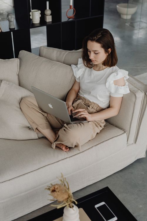 Free A young woman sits comfortably on a sofa using a laptop, capturing a cozy work-from-home setting. Stock Photo
