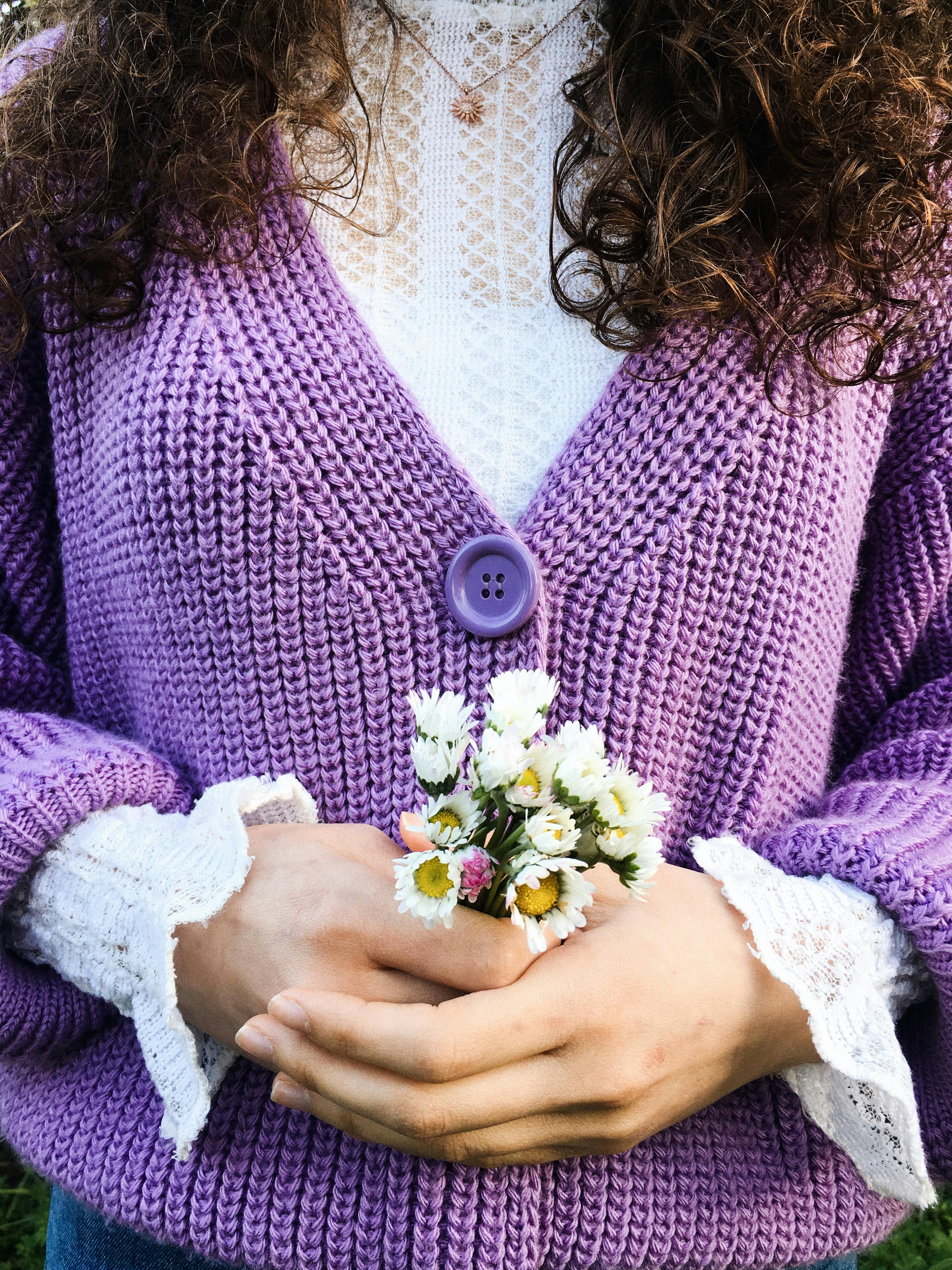 Crop unrecognizable female with curly hair in purple cardigan holding delicate white chamomile on street