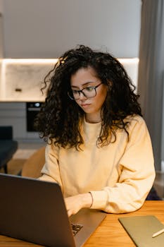 Woman with curly hair in glasses working on a laptop indoors, wearing a yellow sweater.