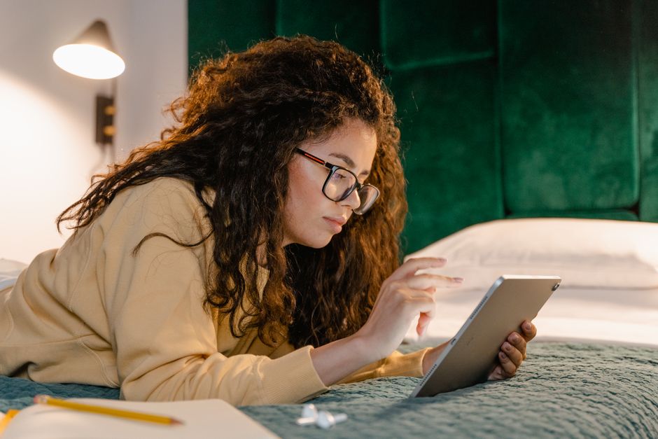 A woman with curly hair working on a tablet in a cozy bedroom setting.