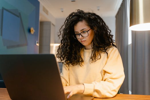Woman in yellow sweater working on a laptop in a cozy home setting.