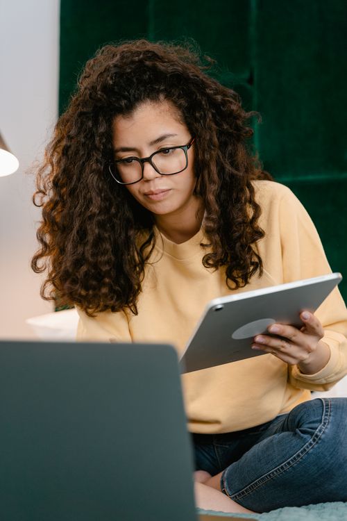 Free Curly-haired woman using a tablet and laptop at home for remote work. Stock Photo