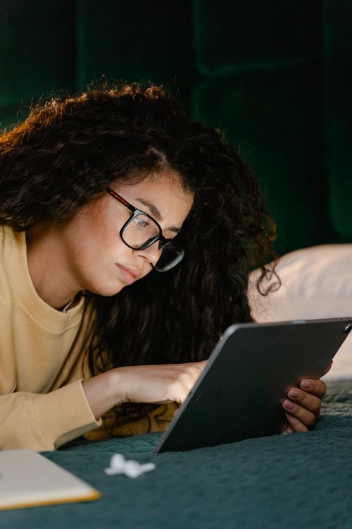 Free Young woman using a tablet on her bed for remote work, wearing eyeglasses in an indoor setting. Stock Photo