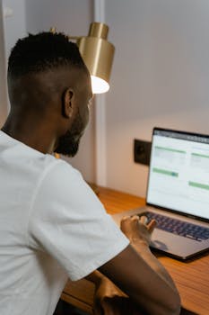 Adult man using laptop at home desk, focused on screen under warm desk lamp light.