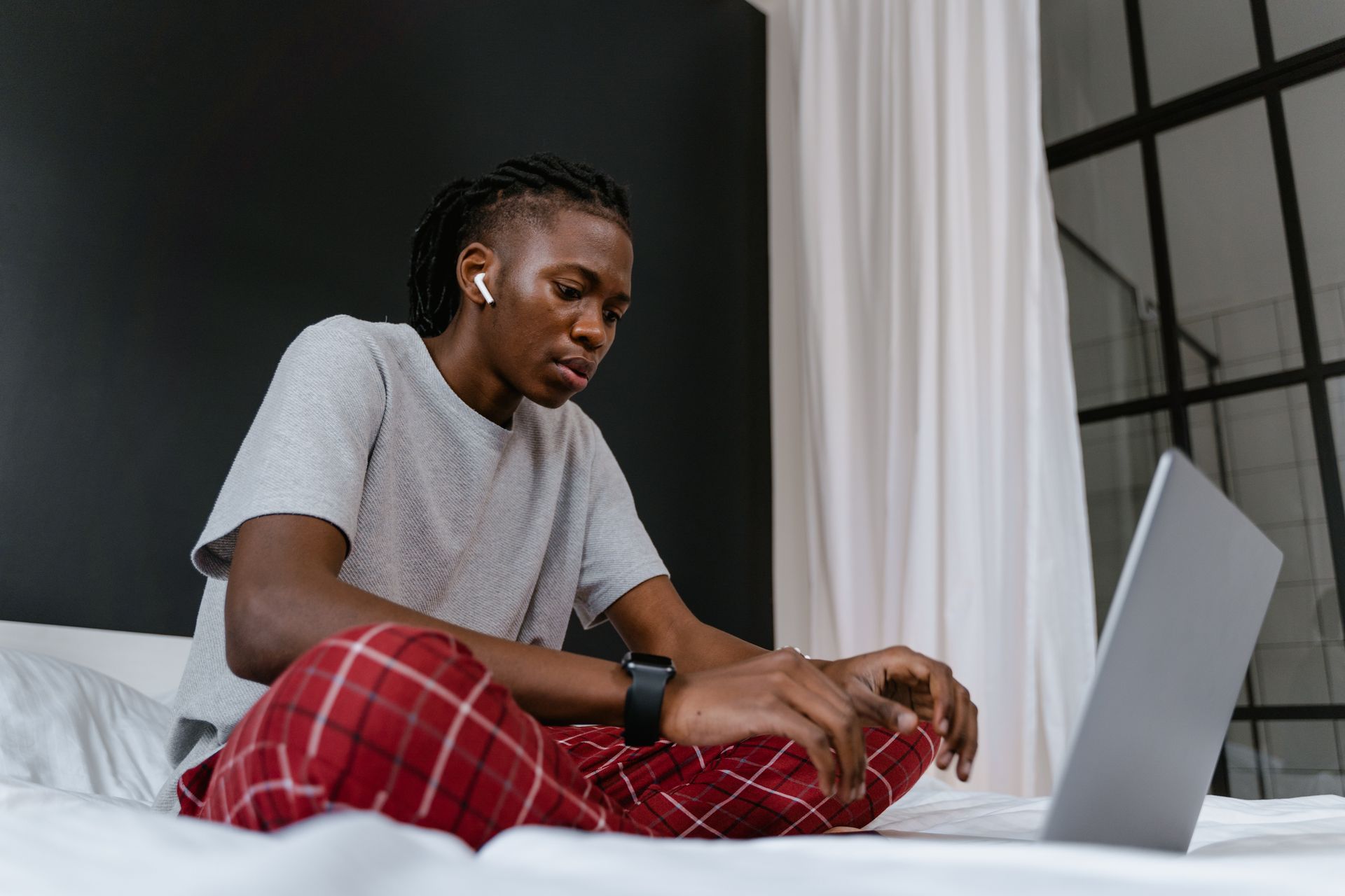 Young adult engaging in remote work from home, sitting on bed with laptop and earphones.