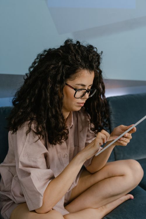 Free Young woman sitting cross-legged on sofa using a tablet, wearing glasses and pajamas. Stock Photo