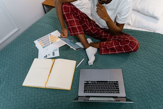 Man in red pajamas working remotely with a laptop and tablet on bed.