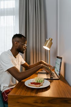 African American man at desk, typing on laptop with a breakfast plate nearby.