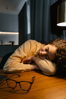 A young woman rests her head on a desk in a dimly lit room, appearing relaxed.