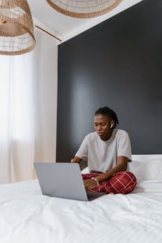 African American man sitting on bed working on laptop, representing remote work lifestyle.