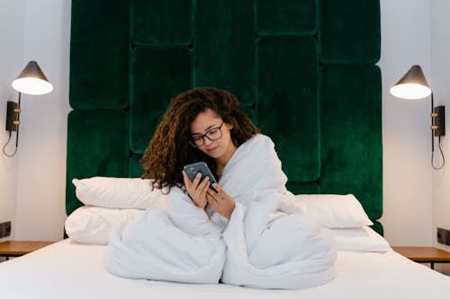 Free A Woman Sitting on the Bed Covered with White Comforter while Using Her Phone Stock Photo