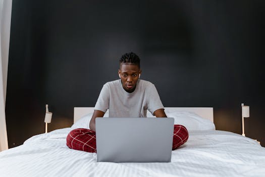Adult man in pajamas using a laptop in a cozy bedroom setting, focusing on remote work tasks.