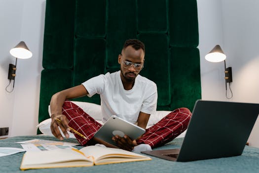 Adult man working from home on a bed with tablet and laptop, wearing pajamas.