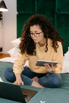 A woman in casual wear works from home using a laptop and tablet, sitting on a bed.