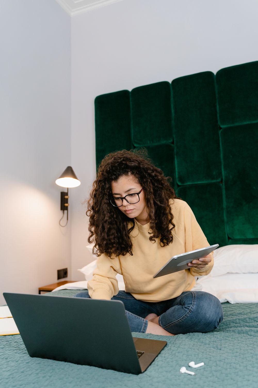 A woman with curly hair and glasses sits cross-legged on a bed, holding a tablet with a laptop open in front of her.