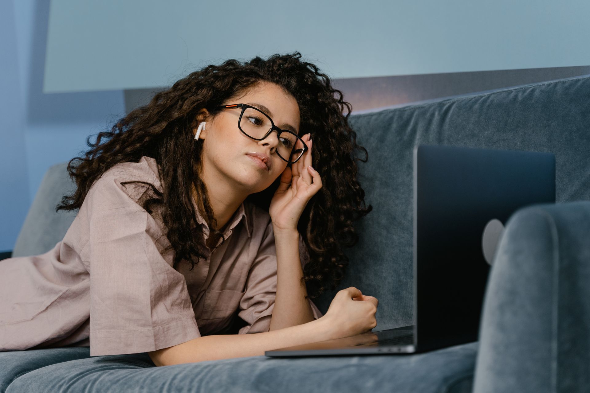 A woman with glasses works leisurely on a laptop while lying on a couch, embracing a remote work lifestyle.