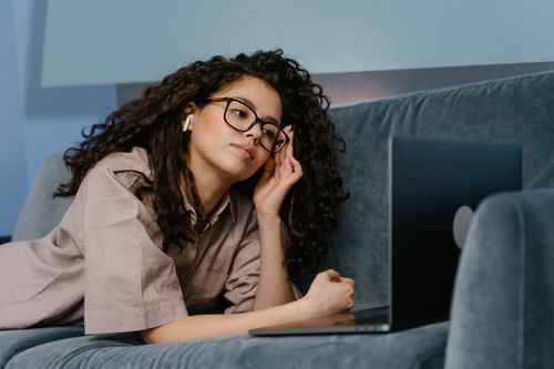 Free A woman with glasses works leisurely on a laptop while lying on a couch, embracing a remote work lifestyle. Stock Photo