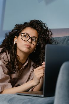 Portrait of a thoughtful young woman with curly hair and eyeglasses using a laptop indoors.
