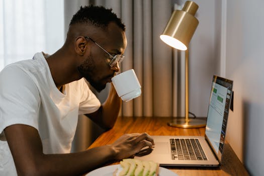 An adult man drinks coffee while working from home on a laptop, creating a cozy workspace vibe.