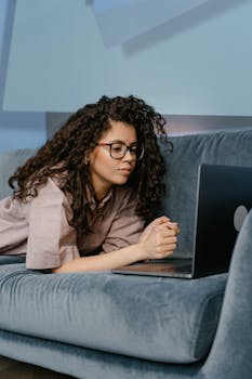 Woman with curly hair and glasses working on a laptop while lying on a sofa.