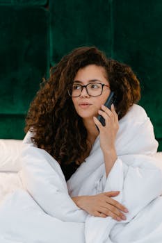 Young woman with curly hair using phone while wrapped in white blanket, relaxed setting.