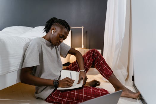 Adult in casual attire working from home with a laptop and notebook.