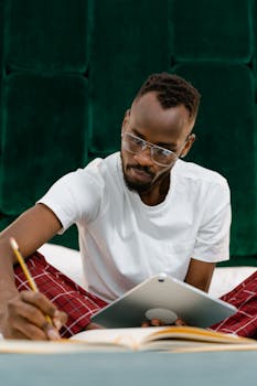 Adult man in glasses writing in a notebook on bed, using a tablet for work from home setup.