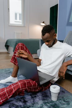 Focused man in pajamas working on laptop at home, seated on the floor.