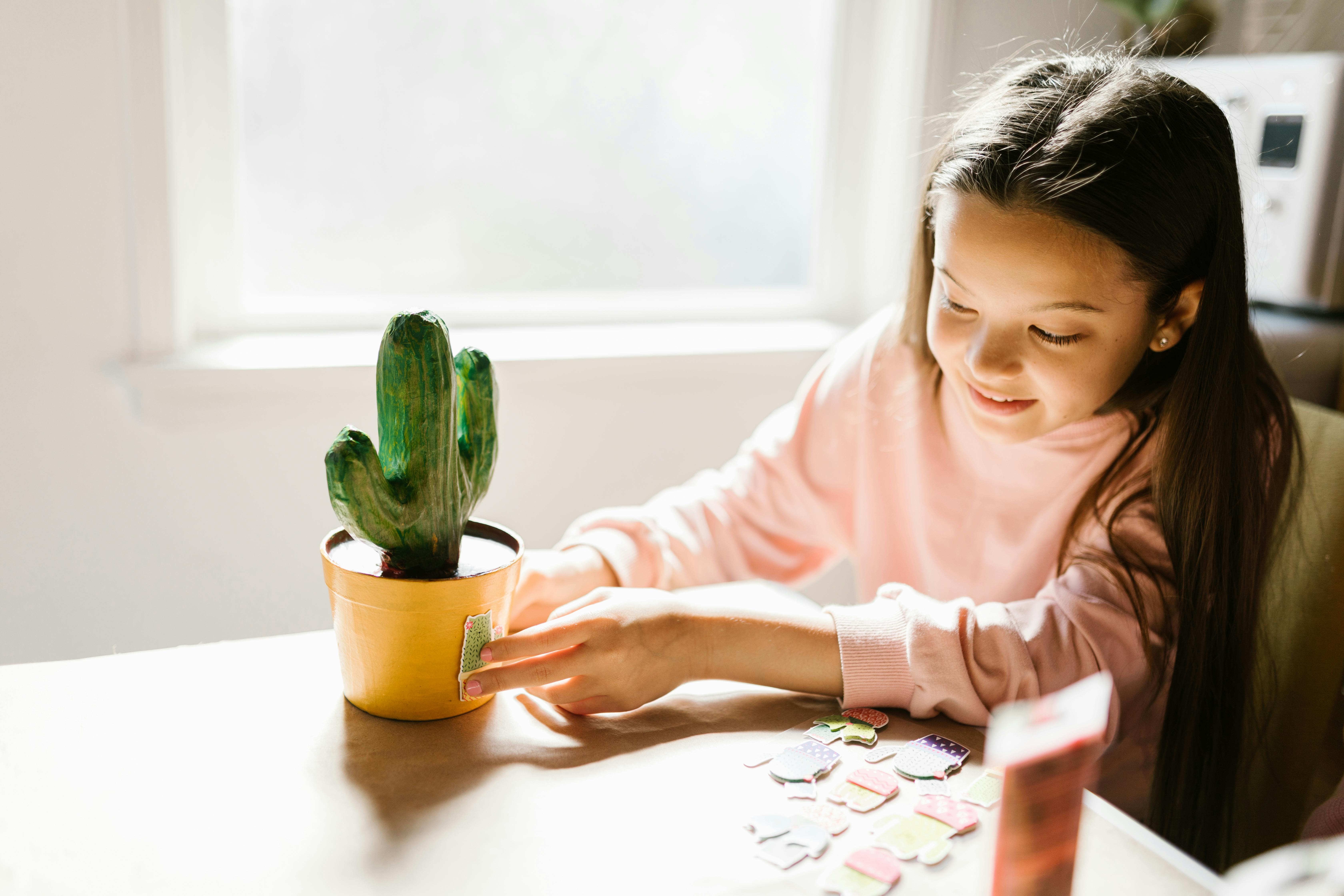 A young girl decorating a cactus pot with stickers in a bright, sunlit room, showcasing creativity and joy.
