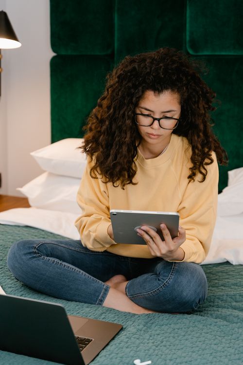 Free Young woman in casual outfit using a tablet while sitting cross-legged on a bed indoors. Stock Photo