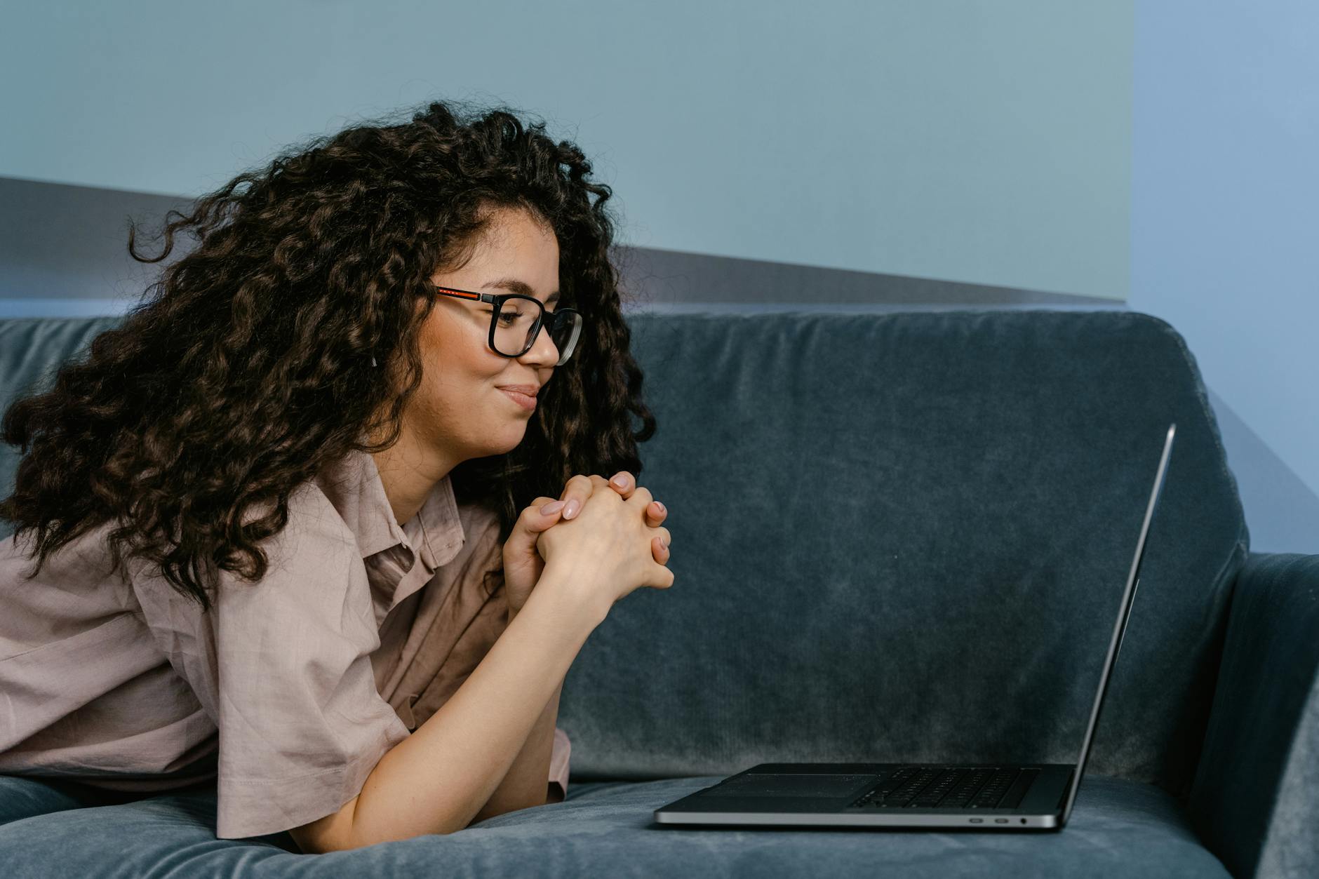 Woman with glasses lying on a sofa working remotely from home on a laptop, relaxed atmosphere.