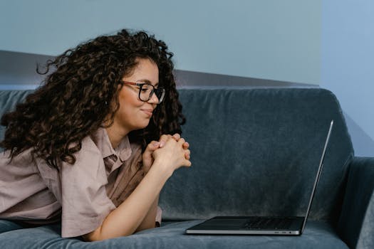 Woman with glasses lying on a sofa working remotely from home on a laptop, relaxed atmosphere.