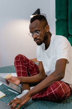Man in pajamas working on a tablet from home, sitting on a bed.