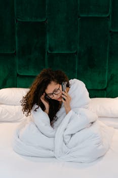 A woman sits wrapped in a comforter, engaged in a phone call, set against a plush green headboard.