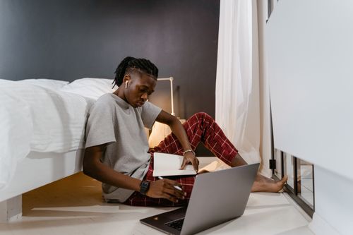 Free A young man in red pajamas works on a laptop and takes notes while sitting beside a bed. Stock Photo