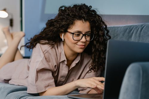 Free Smiling woman with curly hair and eyeglasses working on a laptop indoors. Stock Photo