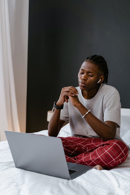 Free Young man sitting on bed, working remotely using laptop in a cozy bedroom environment. Stock Photo