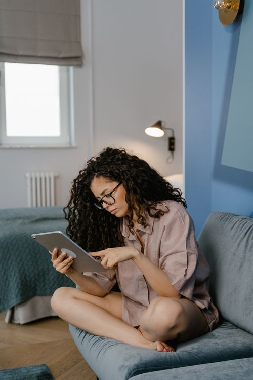 Free Woman with curly hair using a tablet while sitting on a sofa, engaging in remote work or leisure indoors. Stock Photo