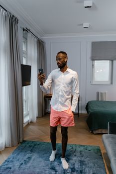 Adult male in smart casual attire using a smartphone for remote work in a bedroom setting.