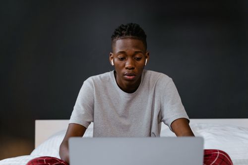 Free Young man focused on laptop screen while working from home, sitting on bed. Stock Photo