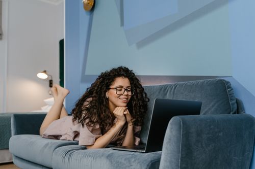 Free Happy woman with curly hair video calling on laptop, lying on sofa in cozy living room. Stock Photo