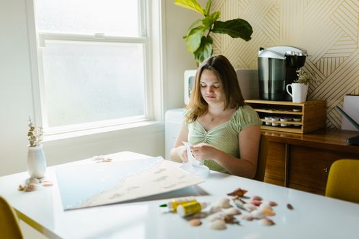 A young woman crafting at a table in a well-lit room with a coffee maker and indoor plants.