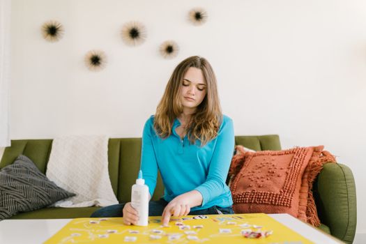 A young woman in a blue sweater sits indoors working on a creative craft project.