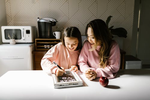 Woman Sitting Beside a Girl Writing on a Board on a Table