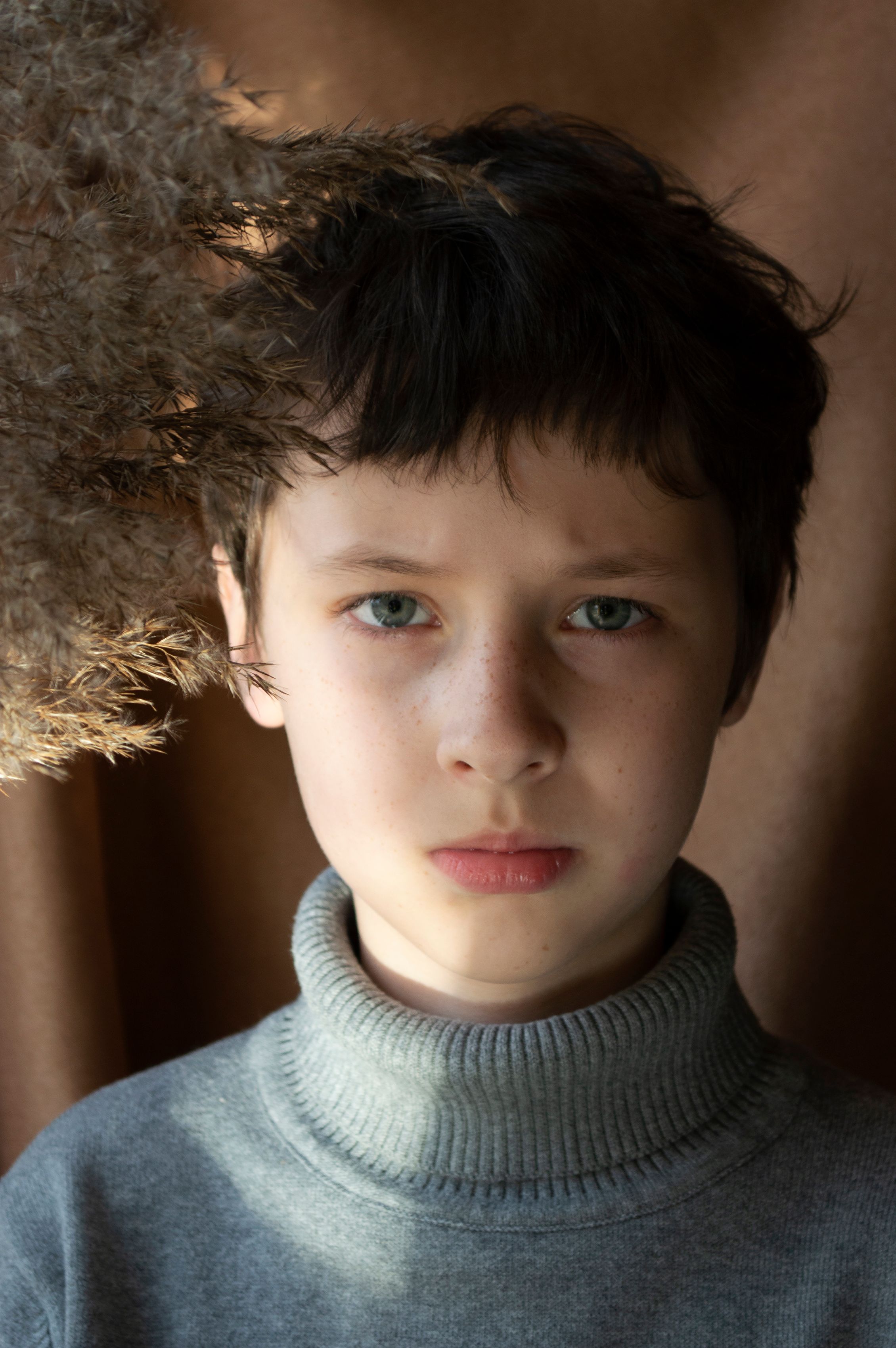 Calm boy with dark hair in gray turtleneck looking at camera with serious face while standing near dried twigs in room