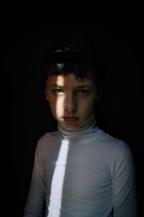 Pensive boy in black hat looking at camera while standing in shadow in dark room with sunbeam on black background