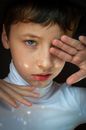 Serious boy in white turtleneck looking away and touching face while covering from blinding sunlight on black background in room