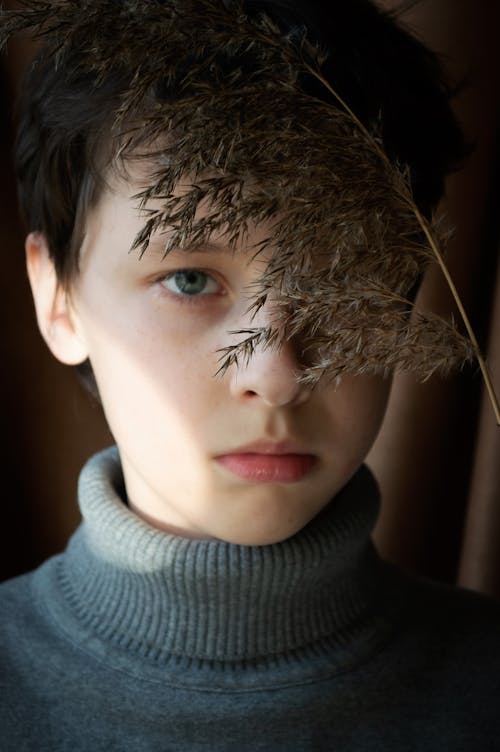Calm boy in warm turtleneck looking at camera with offended gaze near dried plants while standing in sunlight