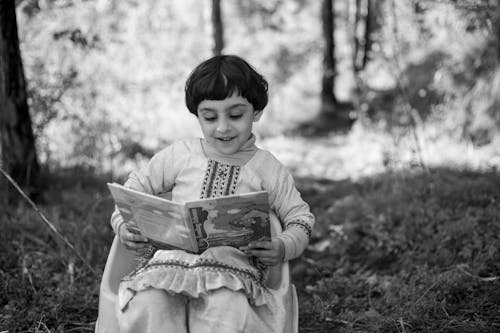 A Grayscale Photo of a Young Girl Sitting while Reading a Book