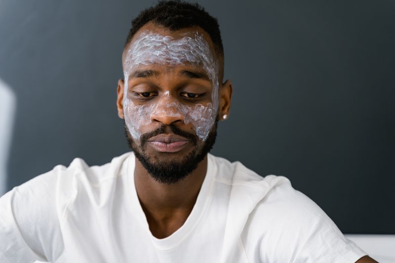 A Black man with deeply pigmented skin and a close-cut beard along his jaw and neck sits in a white t-shirt, looking down, with pale skincare cream spread across his forehead, cheeks and the bridge of his nose.